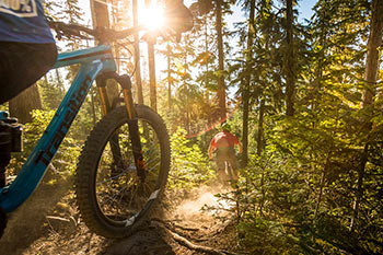 front tire of mtn bike tire with biker in front riding down forested trail with sun light peaking through the trees